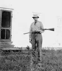picture of a man with a moustache, standing, holding a rifle and wearing a hat
