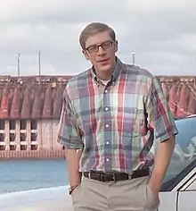 Man with short brown hair and glasses wearing a colorful short-sleeved button-up shirt infront of a bridge above water and red-roofed buildings in the distance