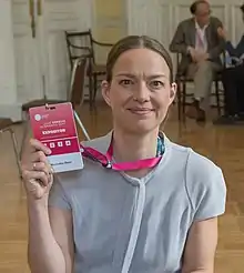 A Caucasian female in a dark blue top, pearl necklace, hair pulled back in a ponytail, holding a microphone.
