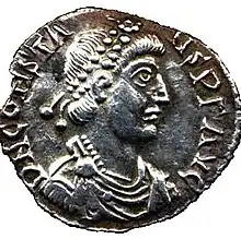 A silver coin showing the profile of a man with two strings of beads in his hair.