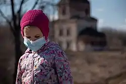 Young girl looking down, wearing a with a pink beanie, floral puffer and a surgical mask.