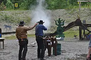 A Cowboy action shooter firing a lever action rifle at steel targets. The Range Officer to the left is holding a shooting timer to measure the time.