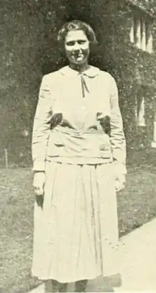 A young white woman standing outdoors, wearing a light-colored dress with peter-pan collar and long sleeves