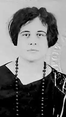 A young white woman with tousled dark cropped hair, wearing a v-neck dark top and dark beads; the edge of an embossed seal is visible in the image