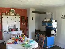 Interior of kitchen: cupboard against wall, table and chairs in foreground, cast-iron stove with pipe leading to chimney