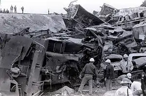 Workers standing around a destroyed freight locomotive, destroyed hopper cars are piled behind the locomotive. Several workers overlook the scene from the railroad track embankment.