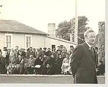 Duncan MacIntyre standing in the foregound at Raukawa Marae in Ōtaki 1971 at a gathering with rows of people sitting in the background and a carved post to one side.