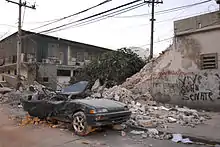 A deep green color car with its roof caved in and the back of the car crushed in. In the background, there is a tree, two phone line poles, and a wall with graffiti on it that is half broken down.
