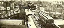 A sepia image of a one-car train with arched windows approaching a three-way junction on an elevated railway with a station on the other side; a side platform exists on the near side of the junction on the track opposite that of the train. There is a wooden car visible at the rightmost track at the station.