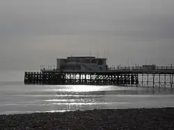 The end of a pier under cloudy skies, illuminated by sunlight reflecting off a calm sea.  There is a small stretch of rocky beach in the foreground.