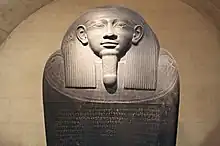 Image of a dark stone Egyptian sarcophagus. The image shows the face of the sarcophagus in a relaxed position looking to the horizon. The sarcophagus shows left-to-right inscriptions in Phoenician on its lid.
