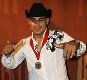 A dark man with black hat, white shirt, raising both hands, with a medal with red ribbon around his neck and legend BMI in the front.