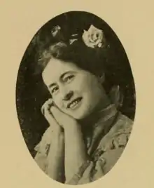 A smiling white woman with her cheek resting on clasped hands, and a rose in her dark hair