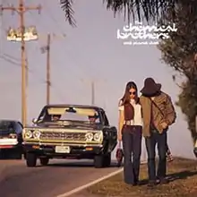 A couple walks down a road on the grass, away from traffic. The man is seen with a large hat which is covering up his head while the woman wears sunglasses while carrying a purse.
