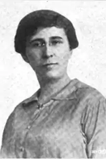 A photograph of a white woman with dark hair; she is wearing eyeglasses and button-down dress or blouse with an open collar