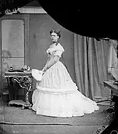 Park, standing in three-quarter profile, wearing a white Edwardian dress and holding a fan