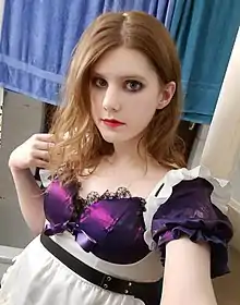 A young male with light brown shoulder-length hair, bright red lipstick, and a maid outfit with purple accents looking into the camera.
