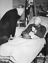 A man lies in a hospital bed, wearing an Army uniform instead of pyjamas. His peaked cap is on the blanket and he holds a baton in his hand. A man in a dark suit and pinstripe trousers bends over to talk to him. In the background are flowers, and a flag.