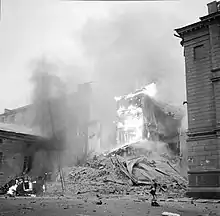 An apartment building is on fire and has partly collapsed in central Helsinki after the Soviet aerial bombing on 30 November 1939. A woman in a coat and a hat is passing on the right next to the rubble, and a car is on fire on the left.