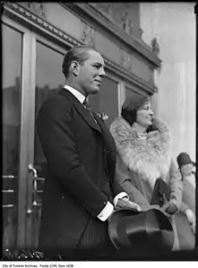 Two people standing in front of two sets of double doors, facing to the right. In front of the nearest set of doors is a man wearing a black suit with striped pants holding a top hat in front of him. In front of the far doors is an older woman wearing a fur-collared coat, clutching a purse in front of her with both hands.