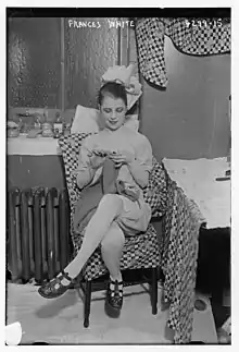 A young white woman wearing a theatrical costume, seated indoors, knitting