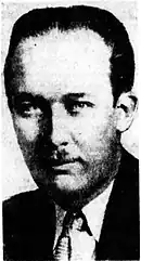 Black-and-white photographic portrait of an expressionless adult man with slicked-back hair, a toothbrush mustache, and a poorly defined cheekbone. His head appears bulbous, being bloated around the forehead and tapering toward the chin. He is depicted wearing a suit.