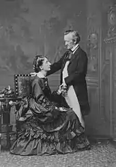 A couple is shown: On the left is a tall woman of about 30. She wears a voluminous dress and is sitting sideways in an upright chair, facing and looking up into the eyes of the man who is on the right. He is about 60, quite short, balding at the temples. He is dressed in a suit with tailcoat and wears a cravat. He faces and looks down at the woman. His hand rests on the back of the chair.
