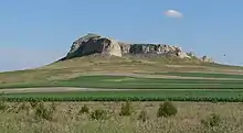 Steep-sided rocky bluff surrounded by grassland; green fields in foreground