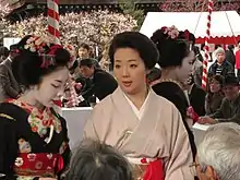 A geisha wearing a plain pink kimono with no white face makeup stood to the right of a maiko in full makeup wearing a heavily-decorated black kimono.