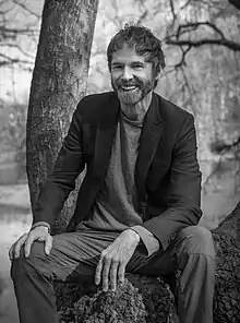 Black-and-white photo of middle-aged white male sitting outdoors by a tree, grinning at camera