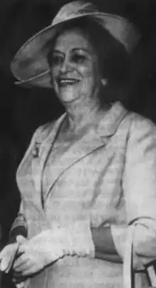 A smiling older Mexican woman wearing a light-colored suit and matching wide-brimmed hat, and gloves
