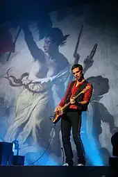 A short-haired man wearing a red shirt and a black waistcoat plays the bass as a picture of Eugène Delacroix's Liberty Guiding the People appears in the background