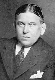 A photographic portrait of critic H. L. Mencken. His hair is parted in the middle, and he appears to be leaning on his left arm. He is wearing a dark tie and a dark suit with peak lapels. A white hankerchief is visible in his suit pocket.