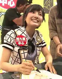 Haruka Kodama in her stage outfit, signing a poster of herself at a HKT48 handshake event in Hong Kong, with two staff members behind her.