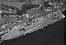 Aerial photograph taken from directly overhead of a small aircraft carrier, while she is berthed alongside a wharf. Nine aircraft are parked in a three-by-three pattern at the carrier's stern, and personnel are positioned to spell out the word "Aloha" when seen from above.