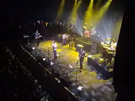 Pop group performing on stage, spotlit, with row of darkened fans in the foreground