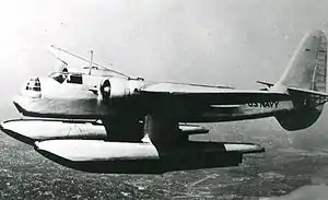 A twin-engined, monoplane aircraft, fitted with two large floats and with a large tail fin, flies over a city.