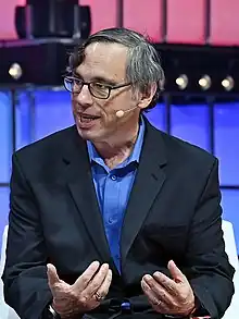 Harry McCracken sitting on a stage in a suit, gesturing with his hands while speaking