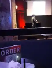 Photo of a sign containing a QR code on a table, with a performer on stage in the background