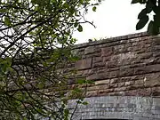 The bridge from the road, looking up through foliage at the east parapet wall with its unornamented date stone.