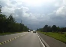 "A rural paved highway running alongside forests and fields. The road is moderately busy. A storm is approaching in the distance."