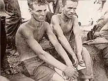 Two men without shirts on sit surrounded by soldiers