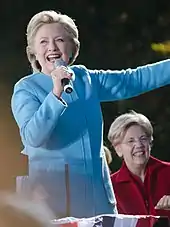  Photograph of Clinton in a light blue suit, holding a microphone and speaking in front of Elizabeth Warren who is seated behind her
