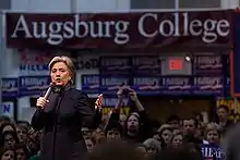 Clinton speaking at a college rally as part of her 2008 presidential campaign, with a crowd behind her looking on. She is speaking at Augsburg College in Minneapolis, two days before "Super Tuesday", the day in 2008 when the largest number of simultaneous state-level elections was held. She is wearing a black suit. There are blue banners with the word "Hillary" on them, hung around the room, as well as a large white-on-burgundy banner with the words "Augsburg College".
