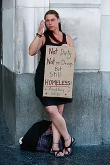 A woman in a black dress and sandals leaning against a building corner holding up part of a cardboard box with "Not Dirty, Not on Drugs, But still Homeless ... Anything helps!" written on it