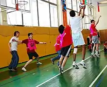 High school or university aged men playing netball on an indoor court. Floor is painted wood. Gym has a window behind the basket.