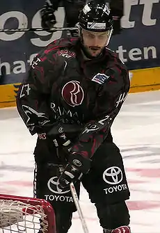 An ice hockey player standing directly in front of the camera. He is wearing a black helmet with a visor and a black and red uniform.