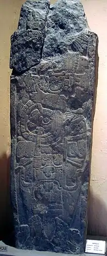 A shaft of stone with a badly damaged upper section. It bears the relief sculpture of a richly dressed human figure, standing and facing left. The head is missing.