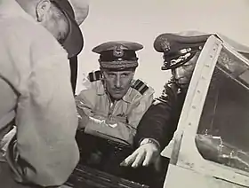 Three men in military uniforms with peaked caps crowding around the open cockpit of a military aircraft, two facing the camera and one with his back turned