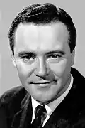 Black and white portrait of Jack Lemmon—a white man with a broad forehead and round face, short dark hair parted on his left, with dark eyes, smiling slightly, and wearing a dark suit, around 43 years of age—in 1968.